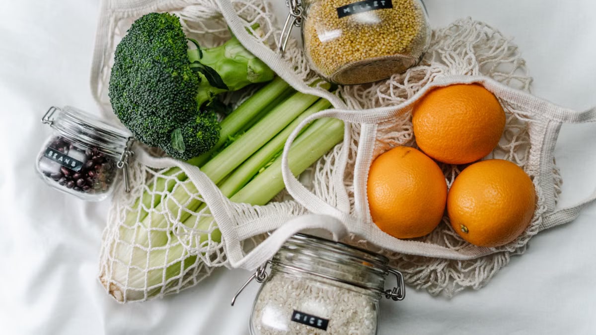 Overhead view of eco bags with broccoli, oranges, and grains in jars, promoting a zero-waste lifestyle.